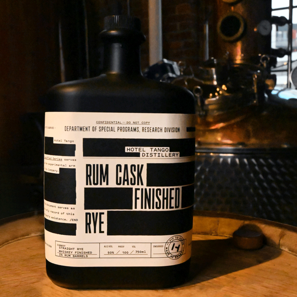 Straight Rye Whiskey Finished in Rum Barrels bottle resting on a rum barrel, with a still in the background.